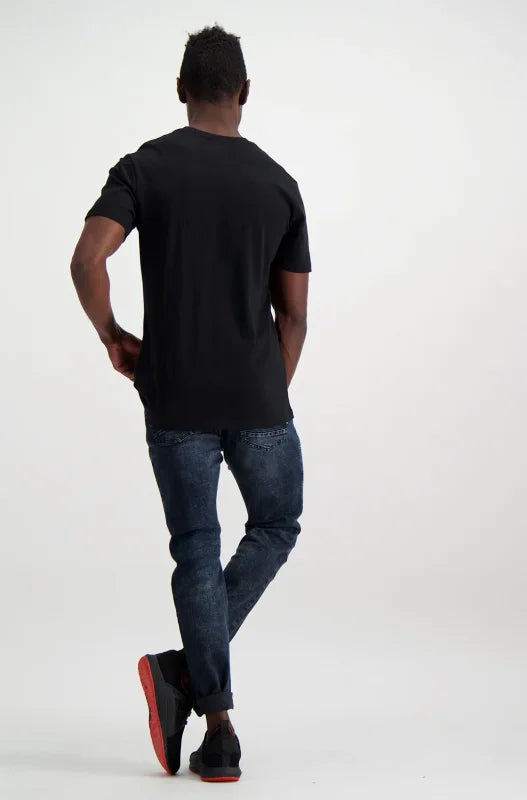 A man modeling a plain black t-shirt, dark blue jeans, and black sneakers with red soles.