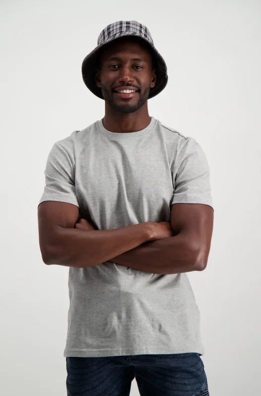 A man wearing a grey t-shirt, dark blue jeans, and a plaid bucket hat.