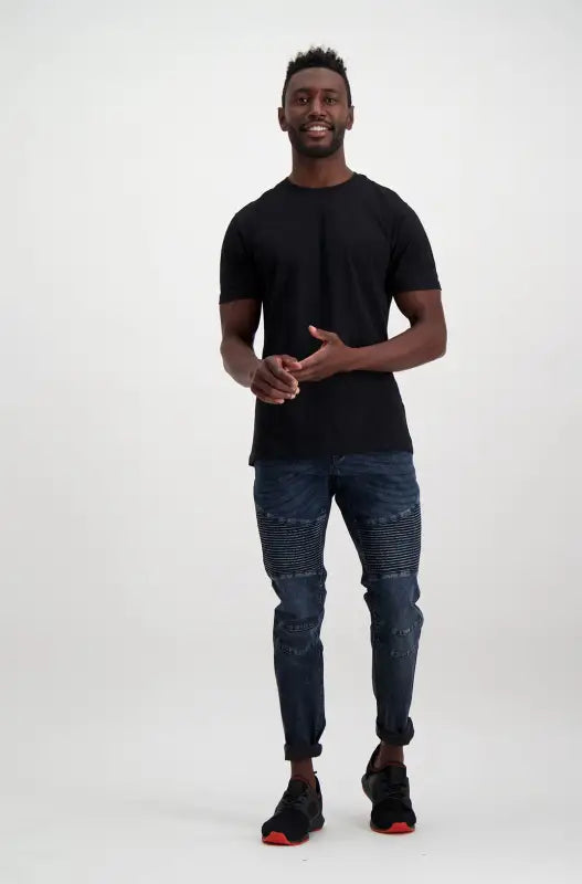 A man in a black t-shirt, dark blue jeans, and black sneakers with red soles