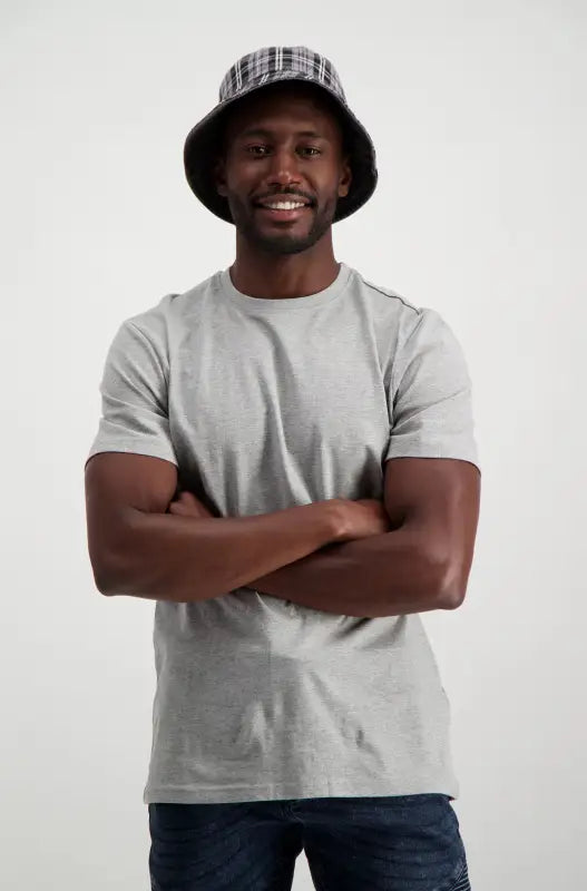 A smiling man in a grey t-shirt, dark jeans, and a black and white plaid bucket hat.