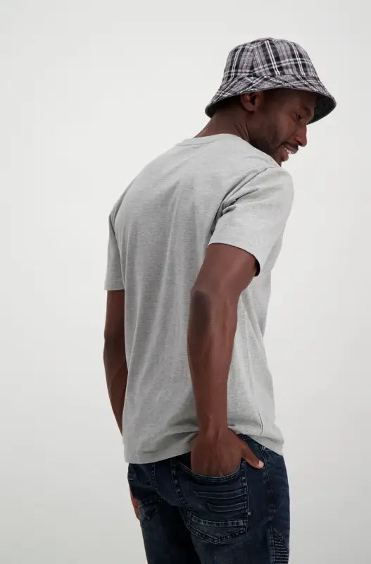 A person wearing a grey short-sleeved t-shirt, dark wash jeans, and a plaid bucket hat.