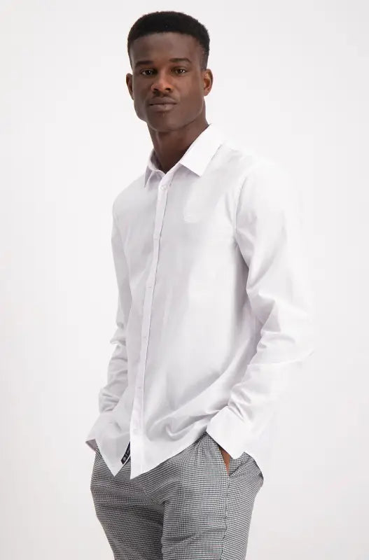 A young man with dark skin, wearing a white long-sleeved button-down shirt and grey and white striped pants, standing with his hands in his pockets.